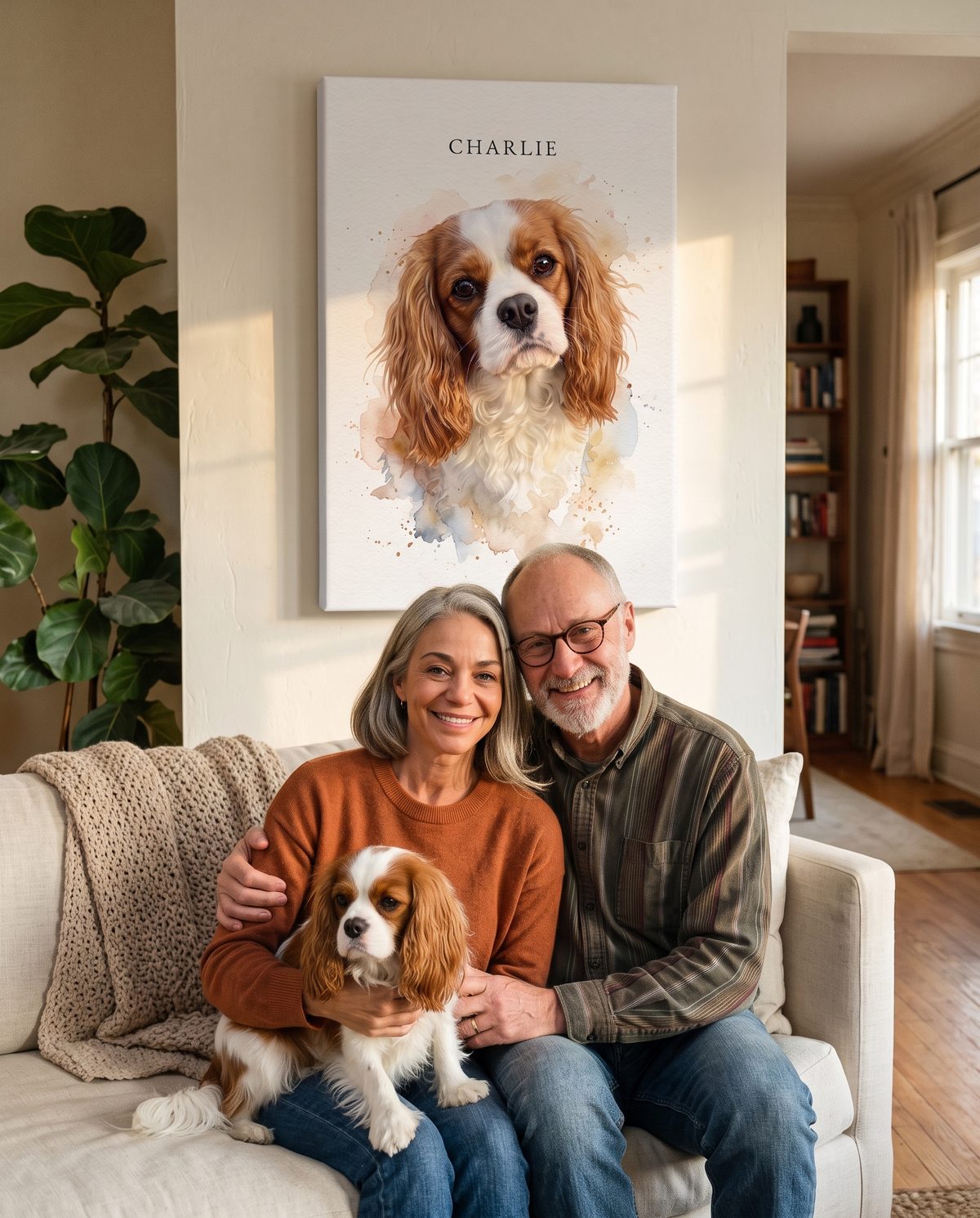 Margaret and her husband Tom on their sofa with their Cavalier King Charles spaniel Charlie, under a tall watercolor canvas portrait of Charlie.